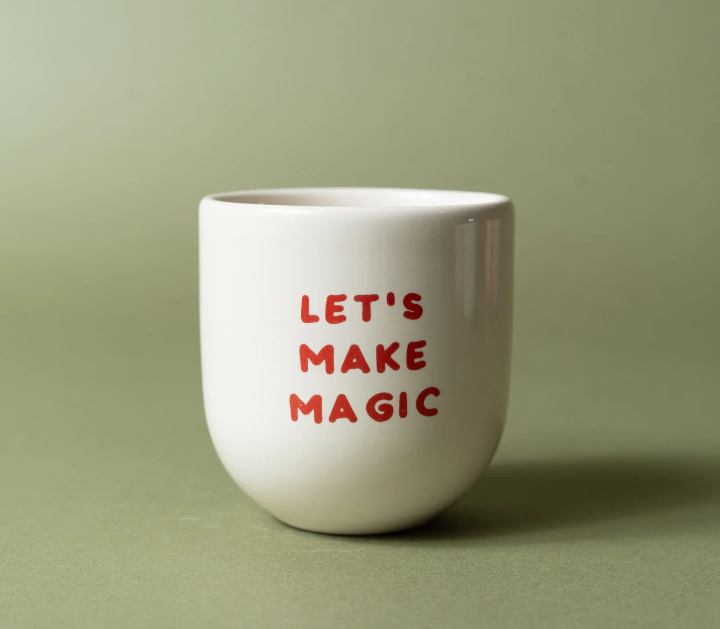 A white ceramic cup with the phrase 'LET'S MAKE MAGIC' printed in red hand-drawn style lettering, set against a solid sage green background.