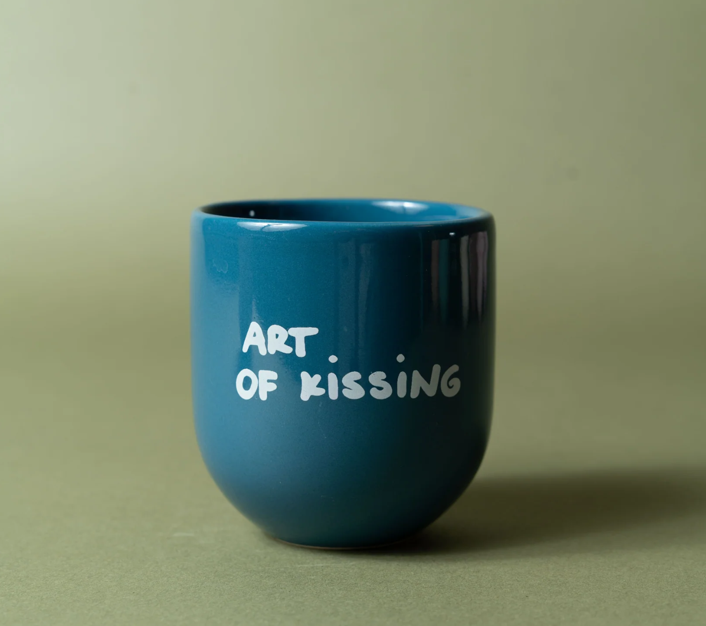A small glossy blue ceramic cup with the words 'ART OF KISSING' printed in white handwritten-style lettering, set against a solid muted olive-green background.