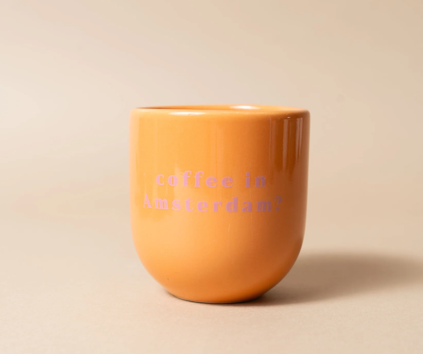 An orange ceramic coffee mug with the text 'coffee in Amsterdam?' printed in pink on the front, set against a plain beige background.