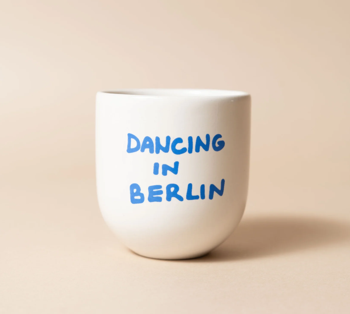 A white ceramic stemless wine glass with the phrase 'DANCING IN BERLIN' printed in blue handwritten-style lettering, set against a solid beige background.