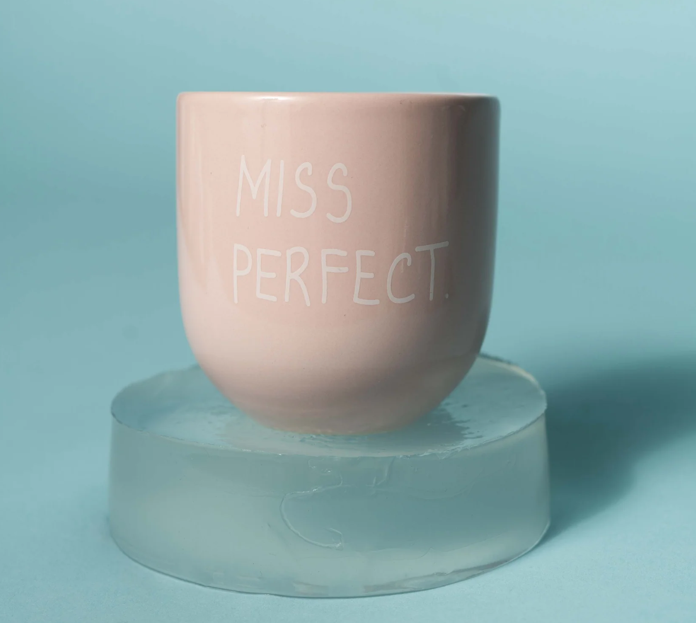 A light pink ceramic cup with the words 'MISS PERFECT.' printed in white on the front, displayed on a translucent acrylic block against a light blue background.
