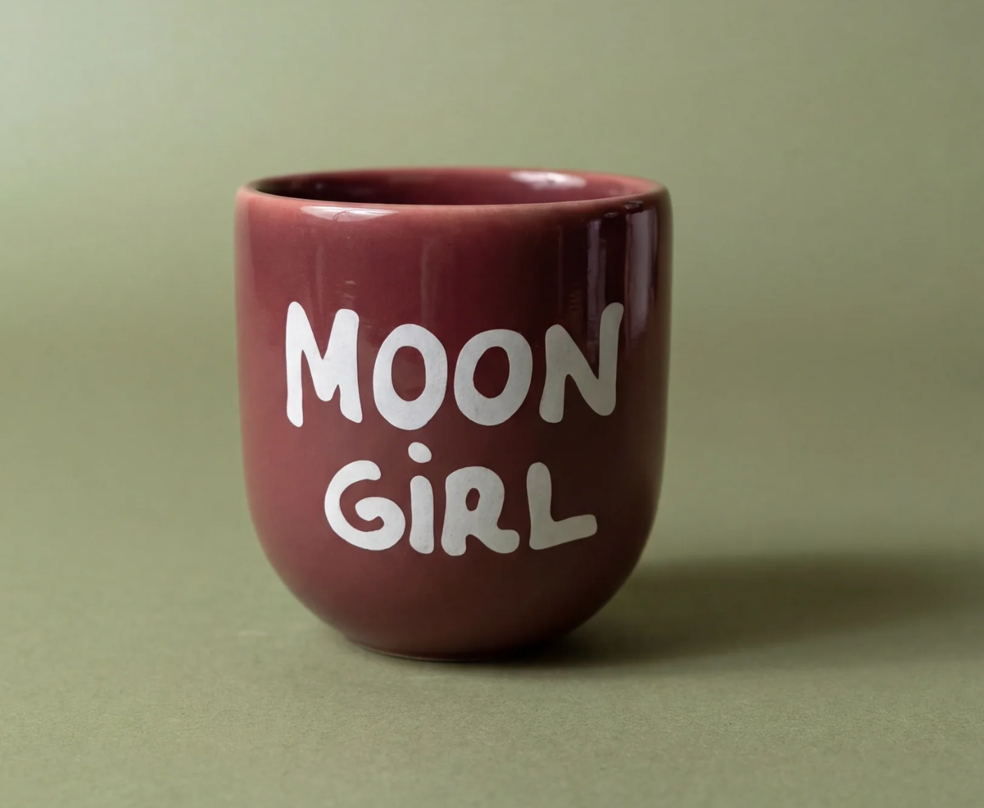A small glossy ceramic cup in a deep burgundy color with the words 'MOON GIRL' printed in white hand-drawn lettering, set against a solid sage green background.