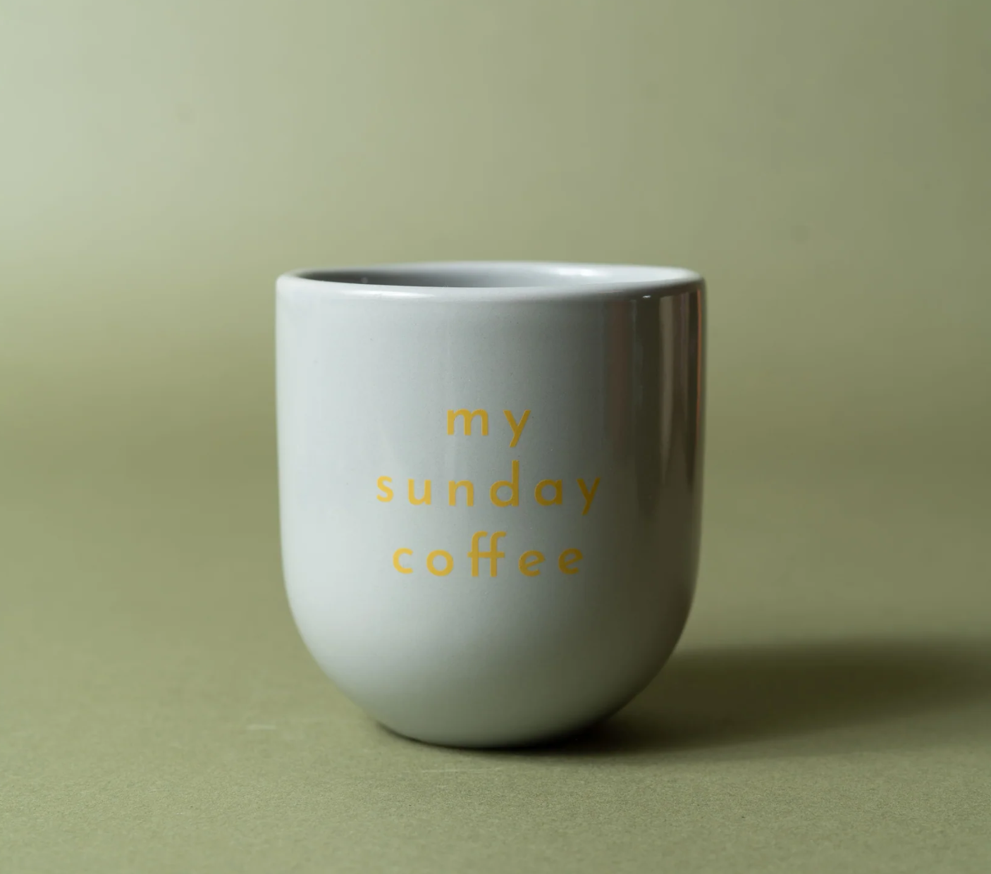 A gray ceramic coffee mug with the phrase 'my sunday coffee' printed in a small yellow sans-serif font, set against a solid olive green background.