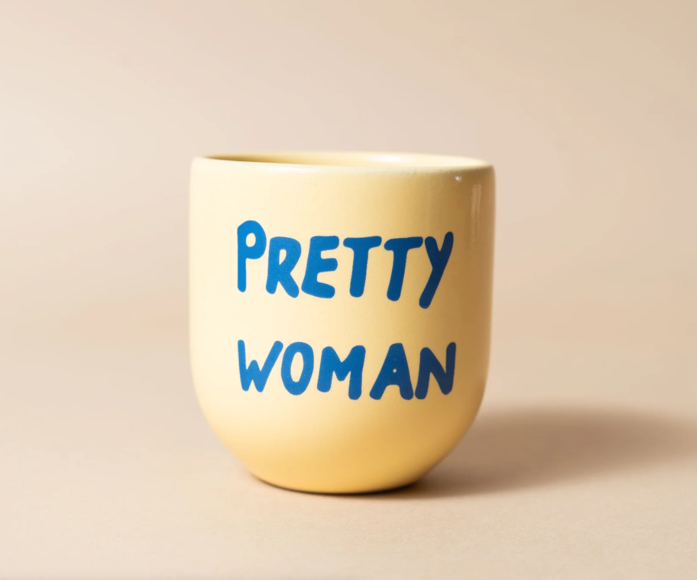 A pale yellow ceramic cup with the phrase 'PRETTY WOMAN' printed in blue lettering, set against a solid beige background.