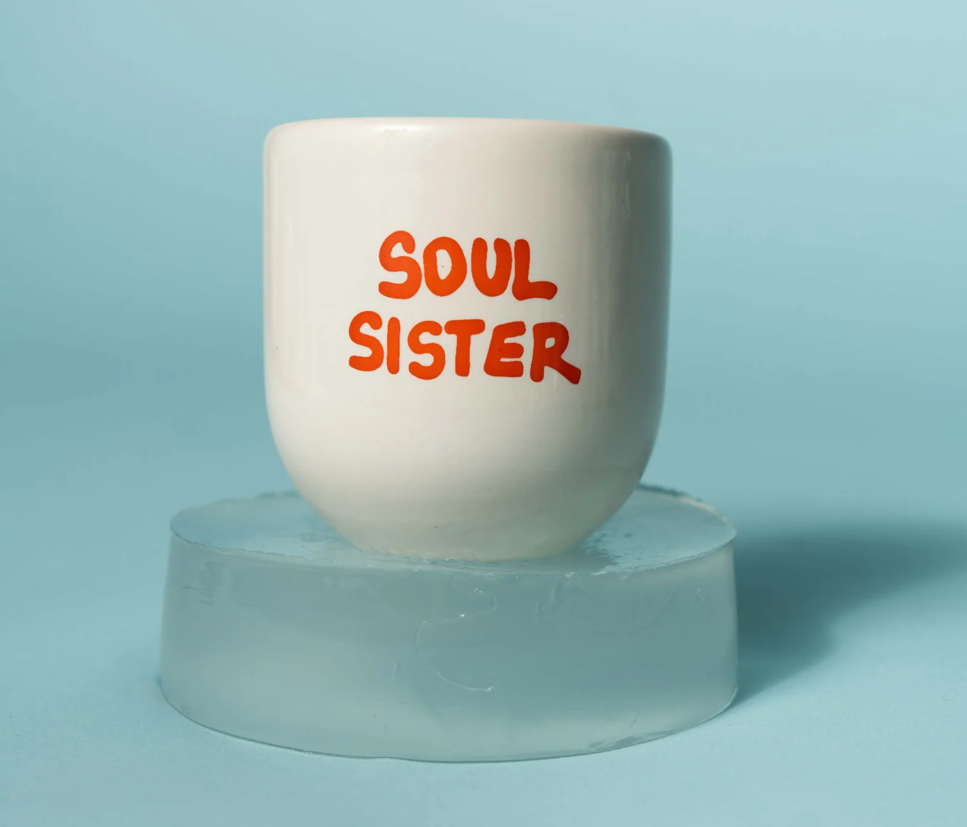 A white ceramic mug with the words 'SOUL SISTER' printed in orange-red lettering, displayed on a translucent ice block against a light blue background.