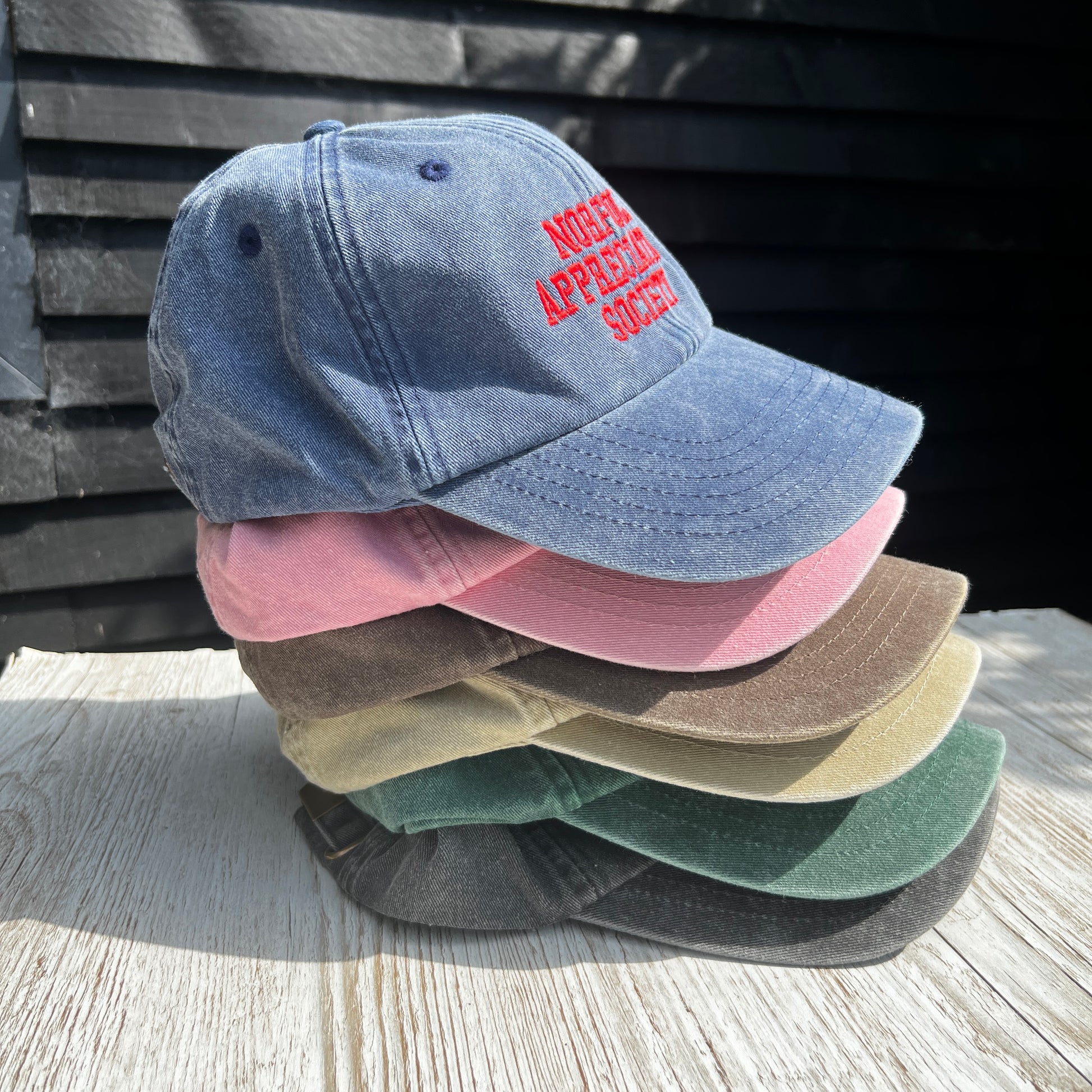 Embroidered 'Norfolk Appreciation Society' caps stacked together in various colours on a light wooden surface