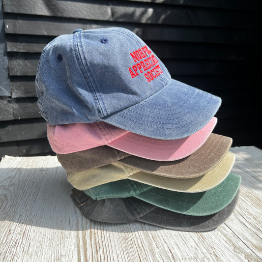 Embroidered 'Norfolk Appreciation Society' caps stacked together in various colours on a light wooden surface
