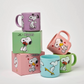 Four pastel coluored Peanuts-themed mugs with snoopy on holding flowers, dancing and skateboarding on a light gray background