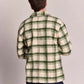 A man viewed from behind wearing a green and cream plaid flannel shirt and dark blue jeans, standing against a plain light gray studio background.
