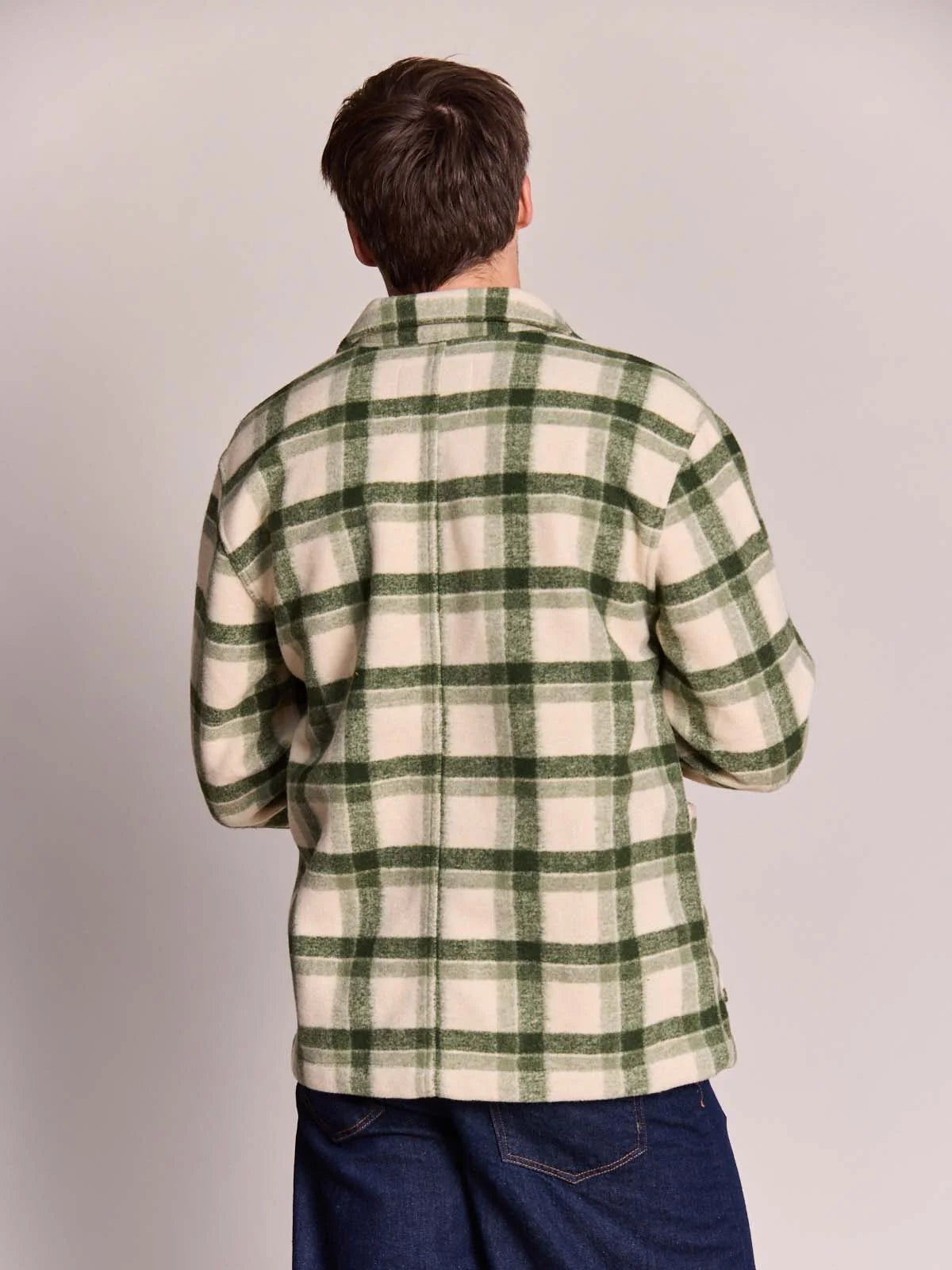 A man viewed from behind wearing a green and cream plaid flannel shirt and dark blue jeans, standing against a plain light gray studio background.