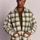 A model wearing a green and cream plaid button-up flannel shirt jacket over an olive green top, photographed against a neutral light gray studio background.