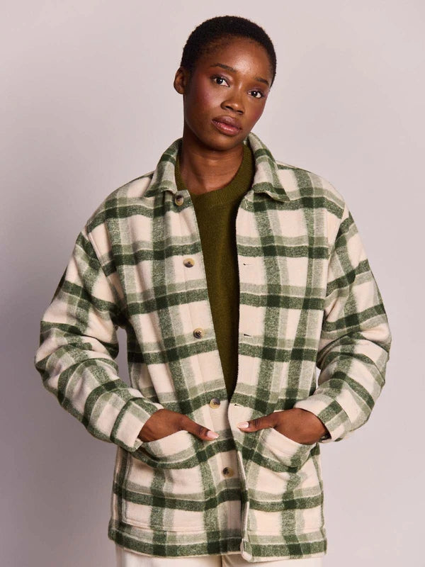 A model wearing a green and cream plaid button-up flannel shirt jacket over an olive green top, photographed against a neutral light gray studio background.
