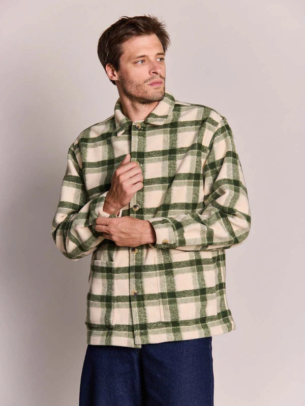 A man wearing a green and cream plaid button-up flannel shirt, paired with dark blue jeans, standing against a solid light gray studio background.